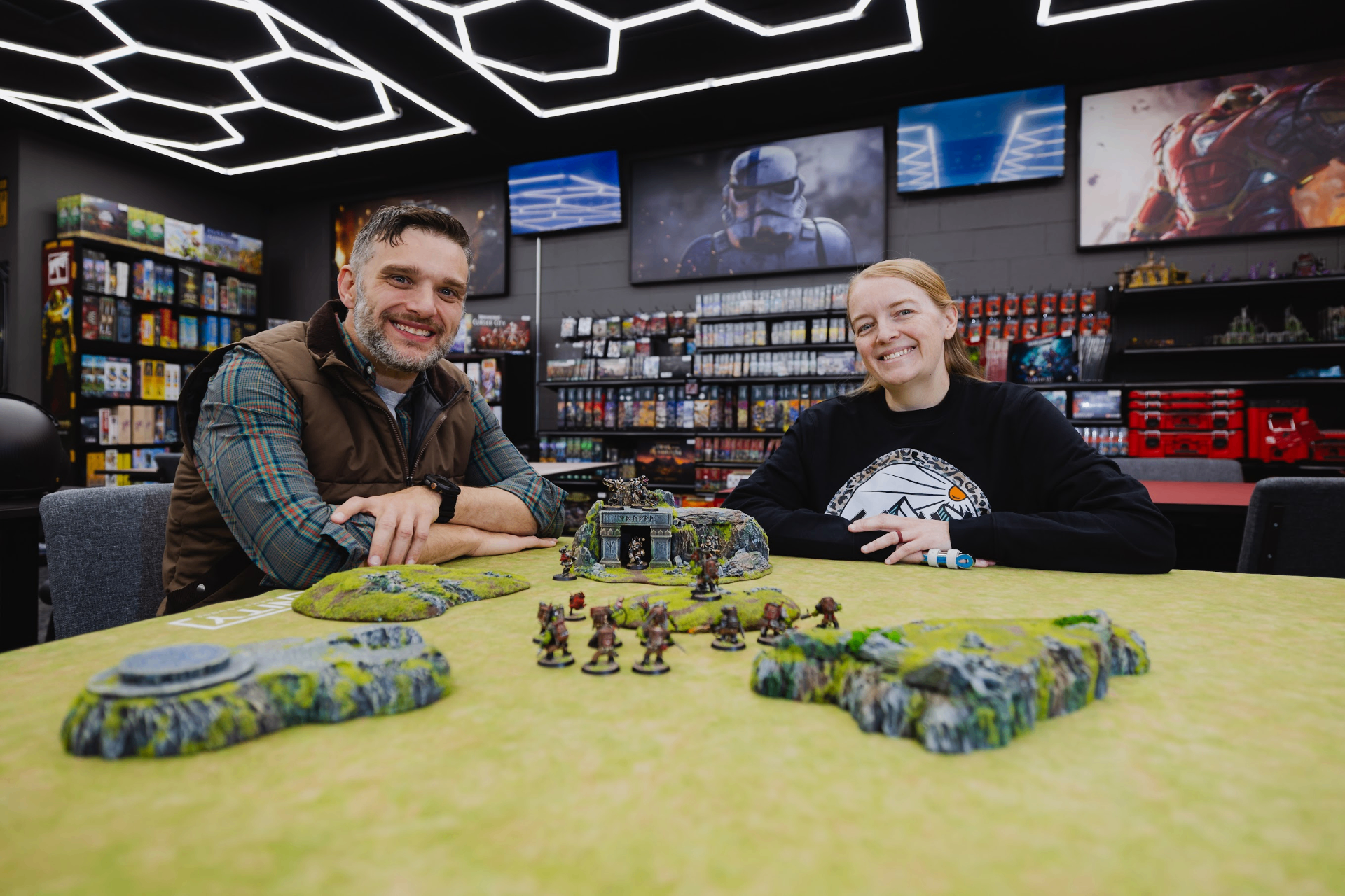 IGL1 store owners smiling at a Warhammer table with painted miniatures and terrain, hex-lit ceiling and backlit shelves of board games behind them.